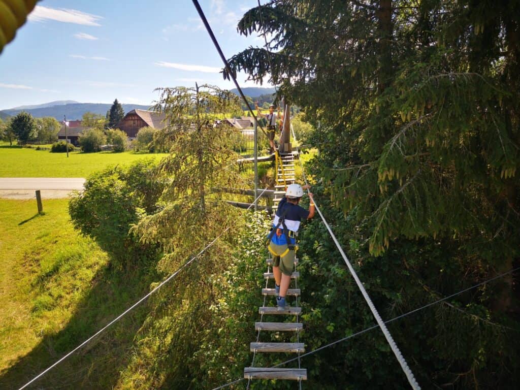 Ein Abenteurer mit Helm und Sicherheitsgeschirr überquert eine schmale Hängebrücke aus Holzplanken, die zwischen hohen Bäumen gespannt ist. Im Hintergrund sind ein grünes Feld, ein Dorf und Berge zu sehen.