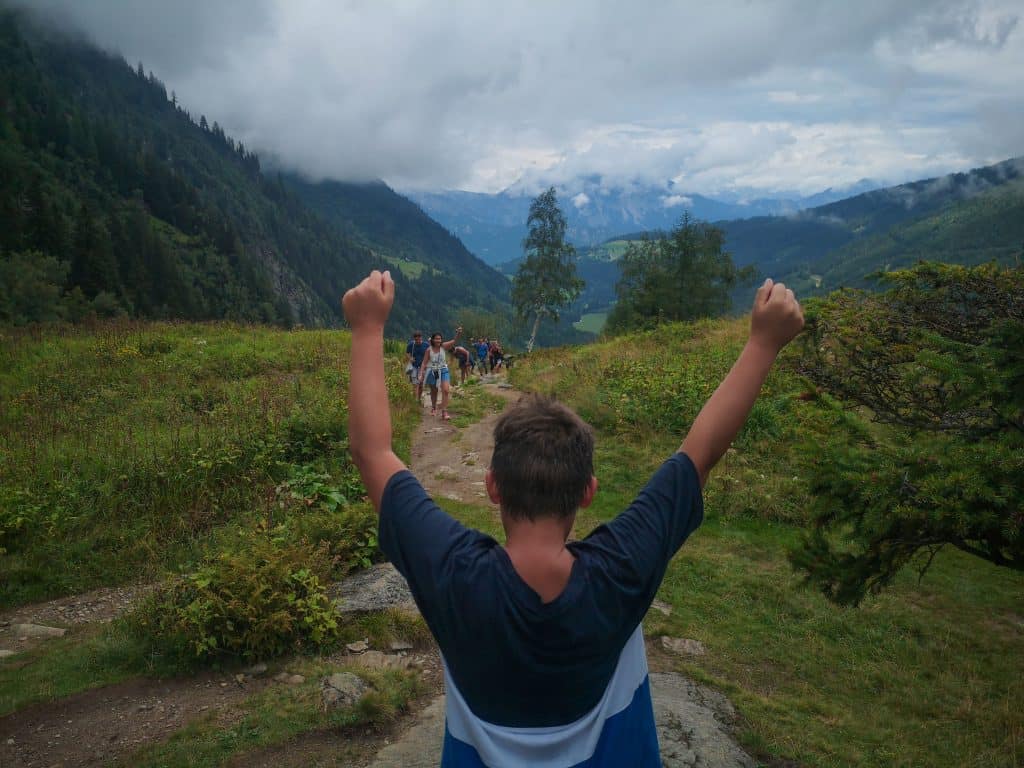 Ein Junge hebt triumphierend die Arme, während er auf einem Wanderweg inmitten einer beeindruckenden Berglandschaft steht. Im Hintergrund ist eine Gruppe von Wanderern zu sehen, umgeben von grünen Wiesen und bewaldeten Hängen, unter einer wolkenverhangenen Himmel.