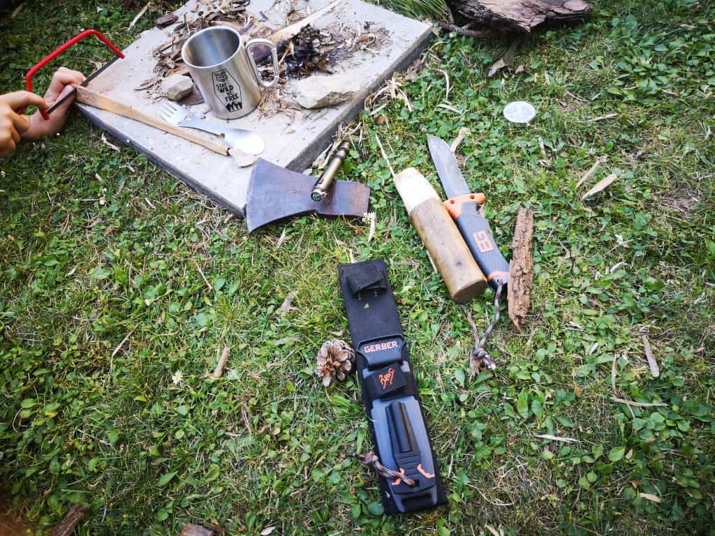 Outdoor-Werkzeuge und Zubehör auf einer Wiese: Eine Axt, ein Messer, ein Holzstück, eine kleine Säge und eine Metalltasse liegen neben einer Feuerstelle auf einer Betonplatte.